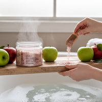 A person's hands are pouring pink bath salt into a glass jar containing apples, with the jar and apples placed on a wooden shelf in a bathroom.