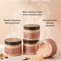 The image showcases a collection of four jars of pink bath salt, each with a black lid, arranged on a marble countertop. The jars are labeled as "New Foaming Bath Salts" and feature descriptions of their benefits, such as "Rich, luxurious foam at an everyday price" and "Deeply hydrating, skin-quenching". The image also includes a small amount of the bath salt spilling out of one of the jars, indicating that the product is in use.