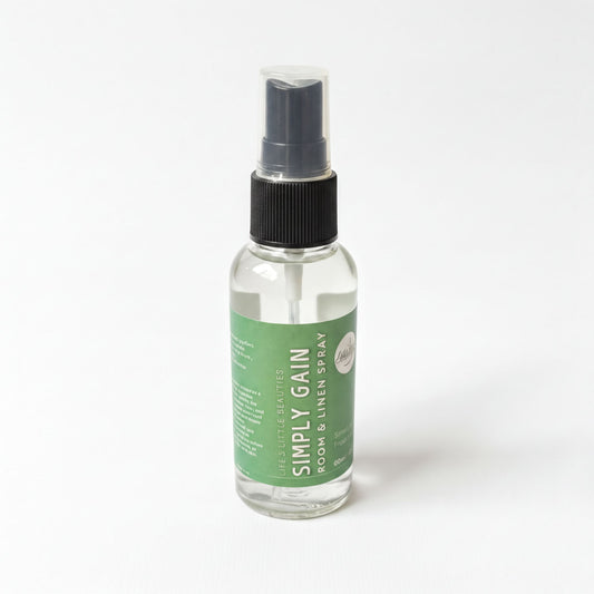 A bottle of "Simply & Gain" spray placed on a stack of white towels on a bed, with a vase of greenery in the background.