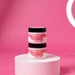 Two jars of pink hair color products are stacked on a white pedestal against a pink background.