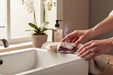 A person's hands are reaching for a bar of soap on a white sink, with a potted plant and a bottle of soap in the background.