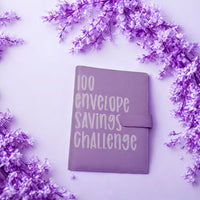 A purple notebook with the text "100 envelope savings challenge" printed on it, surrounded by purple flowers on a light purple background.