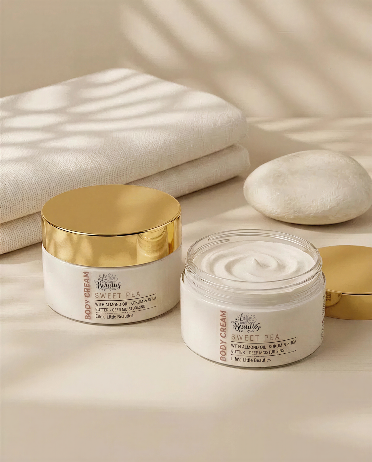 two jars of body cream with gold lids, placed on a white surface. There are also two white rocks and a white towel in the background.