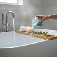 A hand is pouring a blue bath bomb into a white bathtub filled with water, with a lit candle and some greenery nearby.