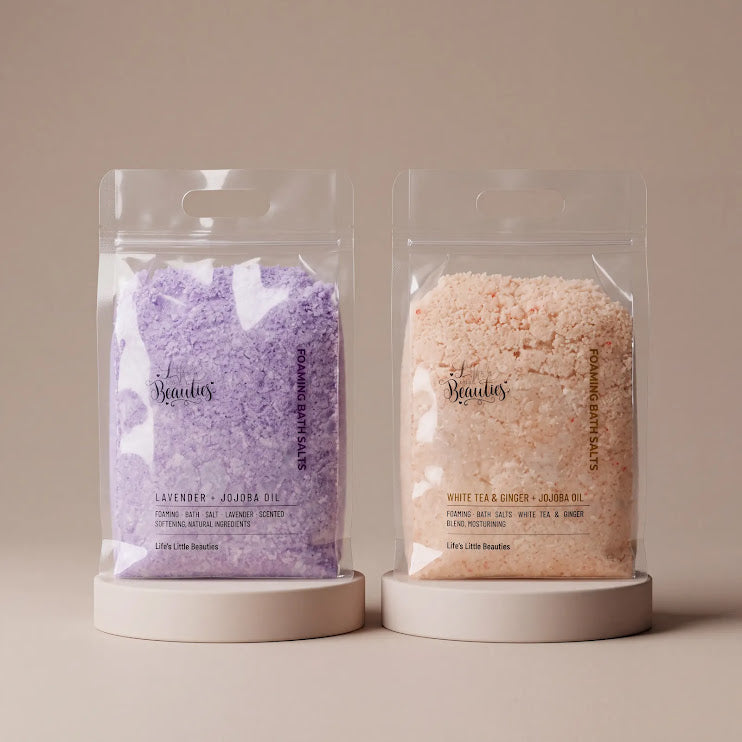 two packages of bath salt, one purple and one beige, placed side by side on a white stand.