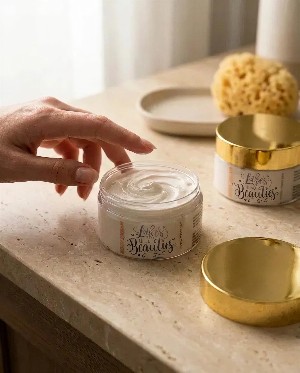 A hand is reaching for a jar of "LESS THAN 500 BeAUTIES" lotion on a marble countertop.