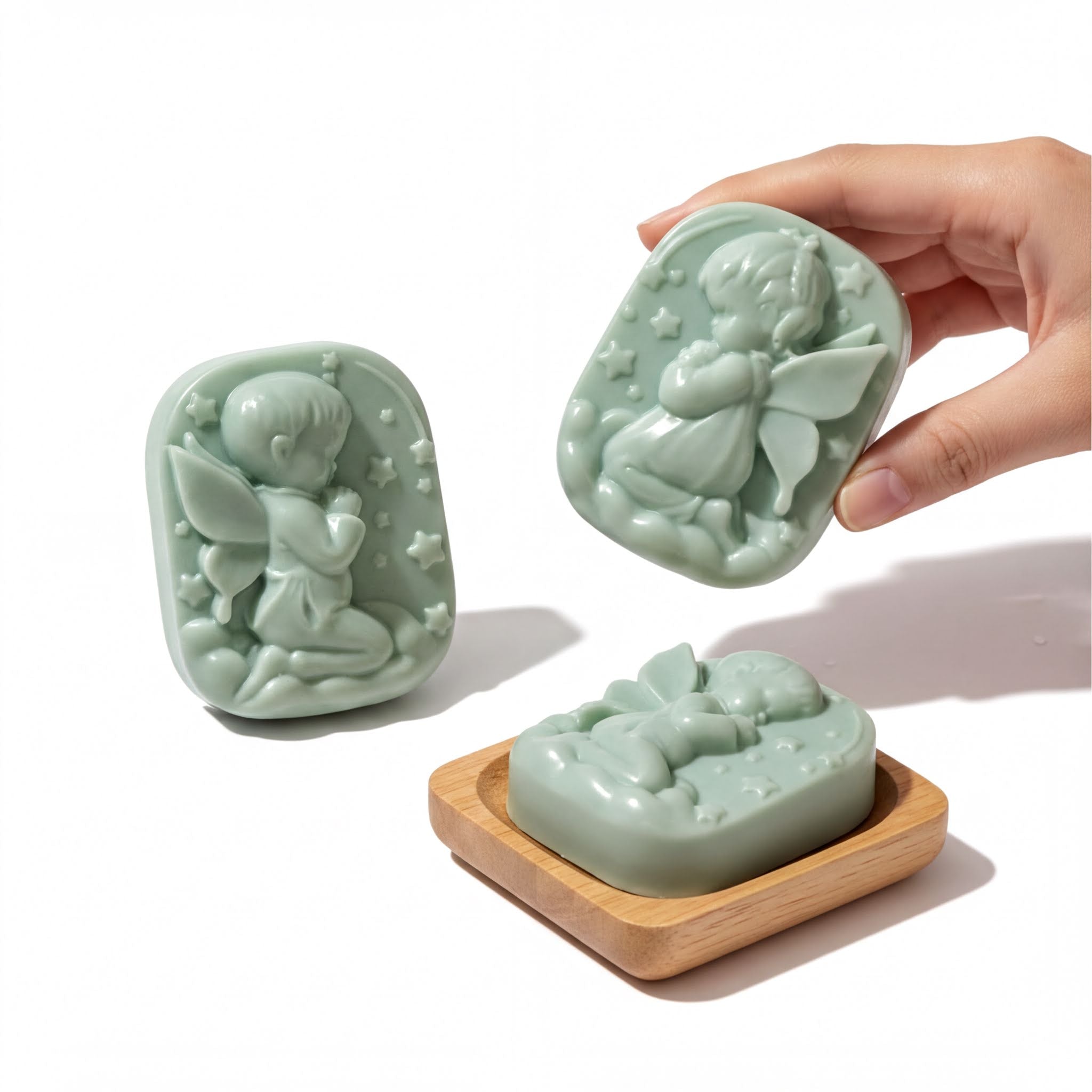A hand is holding a green soap with a fairy or angel design, and there are two other green soap bars with similar designs.