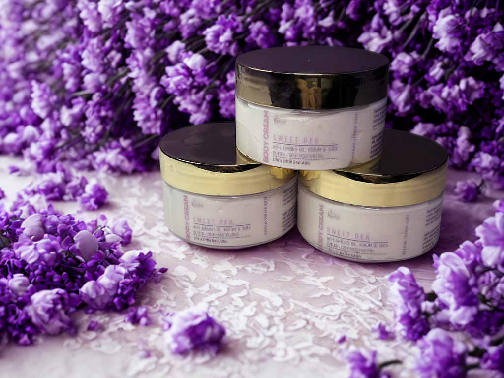 Three jars of "Sweet Pea" body lotion are arranged on a white surface, surrounded by purple flowers.