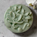 A round, green soap dish with a floral design, featuring a large flower and smaller flowers, is placed on a gray surface.