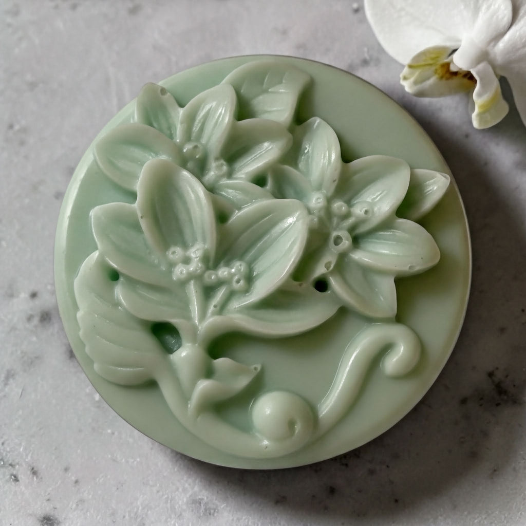 A round, green soap dish with a floral design, featuring a large flower and smaller flowers, is placed on a gray surface.