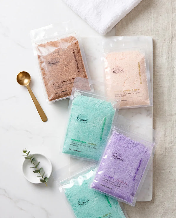 four packages of bath salt, each with a different color, arranged on a white marble surface. A gold spoon is also present in the scene.