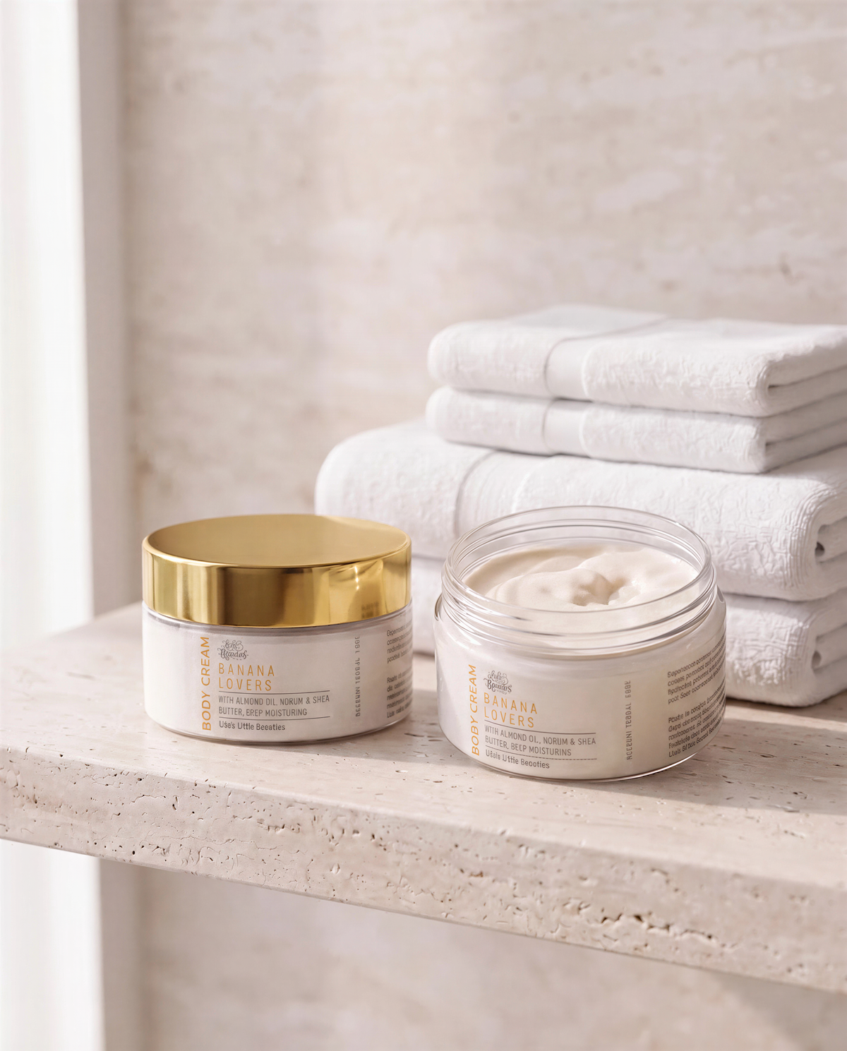 two jars of body cream, one with a gold lid and the other with a clear lid, placed on a white shelf next to folded white towels.