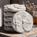 A stack of white, circular soap dishes with intricate floral designs sits on a wooden surface, accompanied by a small glass vase containing dried flowers.